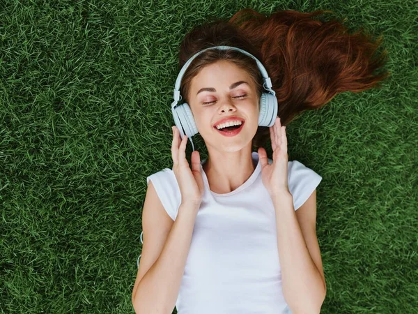 Woman wearing headphones lying on the green grass in a T-shirt and listening to enjoyment with a smile with teeth happiness summer vacations in nature in the park . High quality photo