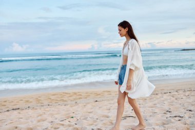 Yüzdükten sonra ıslak saçlarıyla, gün batımından sonra kumsalda kumsalda yürüyen beyaz mayo ve kot şortlu, bronz tenli mutlu kadın. Yüksek kalite fotoğraf