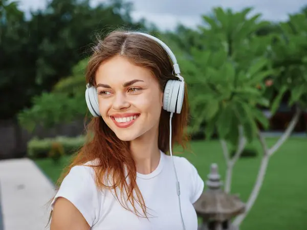 Woman portrait in headphones in a white T-shirt listening to music and walking down the street, smiling, with palm trees in the background. High quality photo