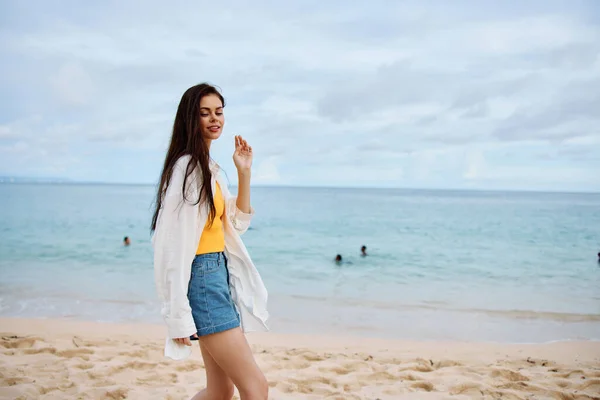 スポーツの女性は 黄色いタンクの上に砂の上の夏の服でビーチに沿って走り デニムショーツの白いシャツは 海の景色 ビーチバケーション 旅行の自由を飛びます 高品質の写真 — ストック写真