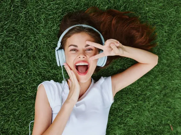 Woman wearing headphones lying on the green grass in a T-shirt and listening to music dancing and singing with a smile with teeth happiness summer vacations in the park . High quality photo