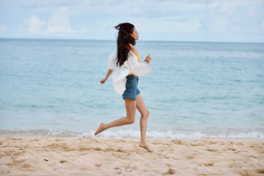Spor kadın plaj boyunca sarı bir atlet ve kot şortla kumsalda koşuyor. Uçan saçlar, deniz manzarası, plaj tatili ve seyahat. Yüksek kalite fotoğraf