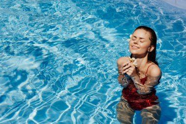 Yüzme havuzunda yüzen, kırmızı mayolu, güzel gülüşlü ve havuzda yüzen Bali çiçekli genç bir kadın. Tatilde rahatlama kavramı. Yüksek kalite fotoğraf