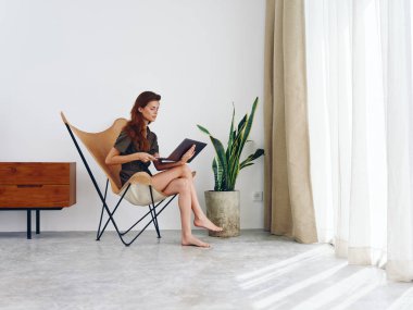 Dizüstü bilgisayarlı bir sandalyede oturan bir kadın film izliyor ve sosyal medyada gülümsüyor, modern stil İskandinav tarzı iç mekan, kopyalama alanı. Yüksek kalite fotoğraf