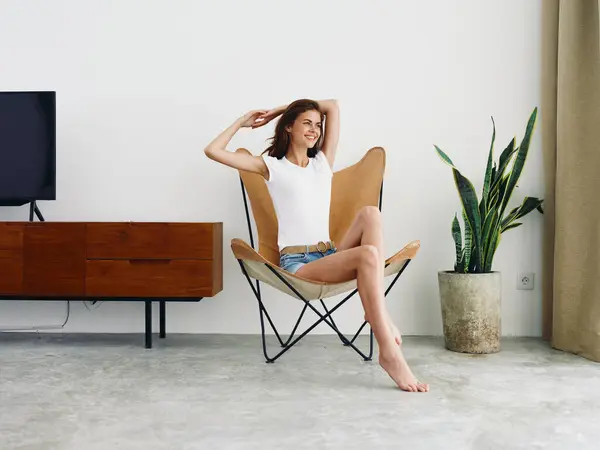 穿着白色T恤和蓝色斜纹棉布短裤的女人坐在皮革扶手椅上笑着 躺在有白色墙壁的时髦的现代室内 复制空间 高质量的照片 — 图库照片