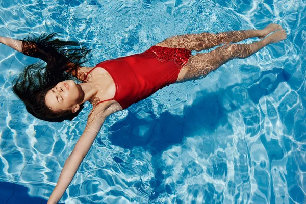 stock image Young woman in the pool in red swimsuit with a beautiful smile lying on the water and swimming in the blue pool in the sun, the concept of relaxing on vacation. High quality photo