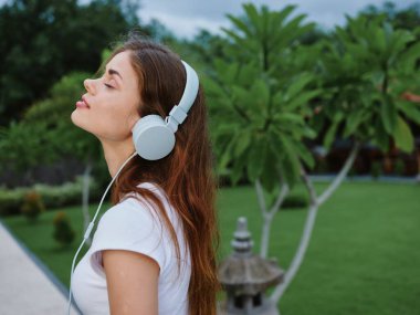 Beyaz tişörtlü, kulaklık takan, müzik dinleyen ve sokakta yürürken palmiye ağaçlarının önünde gülümseyen bir kadın. Yüksek kalite fotoğraf