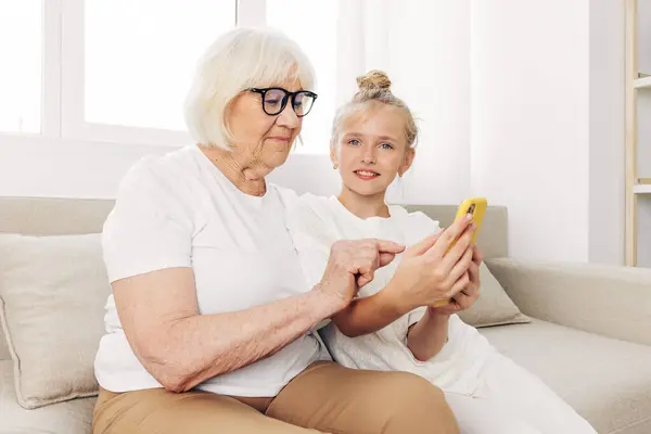 Grandmother family phone indoors child two smiling video selfie education t-shirt granddaughter bonding photography people hugging sofa space copy togetherness call white