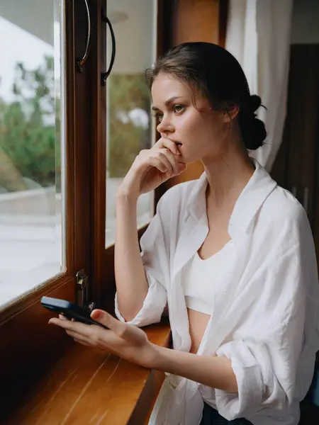 家の窓に手で立っている携帯電話が付いている美しい女性 休暇のフリーランサー オンライン仕事 サイバーセキュリティの概念 スマートな家が付いている家の便利なライフスタイル 高品質の写真 — ストック写真