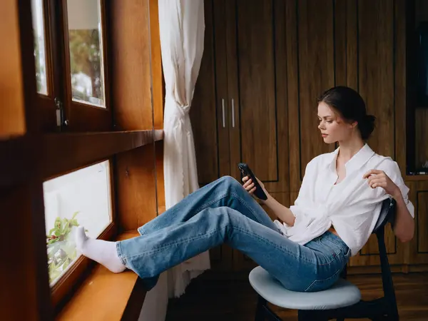 Pencerenin Kenarında Oturan Genç Bir Kadın Elinde Telefonla Gülümsüyor Çevrimiçi — Stok fotoğraf
