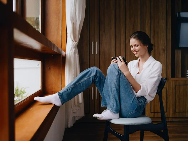 年轻女人坐在窗边 手里拿着电话 微笑着上网看 春天的心情 家里简朴的衣服 家舒适的氛围 美感的生活方式 高质量的照片 — 图库照片