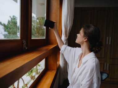 Ev penceresinde elinde telefonla bekleyen güzel bir kadın, tatilde serbest çalışan, çevrimiçi işi olan rahat bir ev hayatı, siber güvenlik konsepti, akıllı bir ev. Yüksek kalite fotoğraf