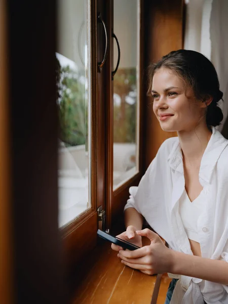 漂亮的女人 手里拿着手机 站在家里的窗口 自由职业者在度假 家里方便的生活方式与在线工作 网络安全的概念 聪明的家 高质量的照片 — 图库照片