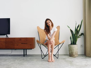 Dişleriyle, beyaz tişörtlü, mavi kot şortlu, deri koltukta oturmuş gülümseyen bir kadın. Beyaz duvarlı, fotokopi odası olan modern bir evde dinleniyor. Yüksek kalite fotoğraf