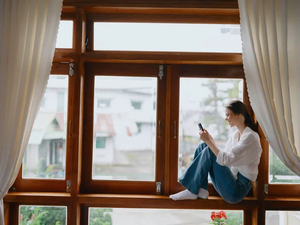 年轻女人坐在窗边的木制窗台上 手里拿着电话 面带微笑地看着窗外 秋天的心情 舒适的家庭环境 美学的生活方式 高质量的照片 — 图库照片
