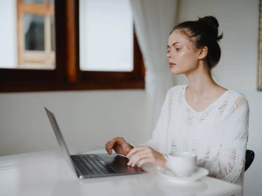 Evde rahat bir yerde, bir bardak içkiyle masa başında çalışan yaratıcı genç bir kadın. İnternette gülümsemesi, iletişimi ve eğitimi olan bir ofis çalışanı. Yüksek kalite fotoğraf