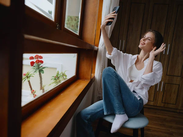 Beautiful woman with phone in hand sitting at the window with wooden frame of the house, home comfortable lifestyle with online work, cozy atmosphere and aesthetics, spring time. High quality photo
