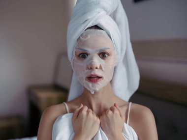 Genç bir kadın, duştan sonra kafasında nemlendirici yüz maskesi ve beyaz havluyla yatakta uzanıyor. Yaşam tarzı, cilt bakımı, gülümseme. Yüksek kalite fotoğraf