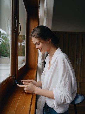 Ev penceresinde elinde telefonla bekleyen güzel bir kadın, tatilde serbest çalışan, çevrimiçi işi olan rahat bir ev hayatı, siber güvenlik konsepti, akıllı bir ev. Yüksek kalite fotoğraf