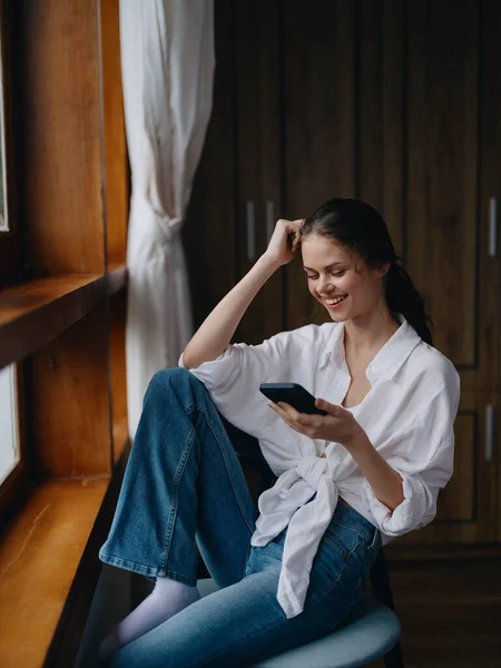 Young woman sitting at the window on a chair with a phone in hand smiling and looking out the window, spring mood, home cozy atmosphere, aesthetic lifestyle. High quality photo