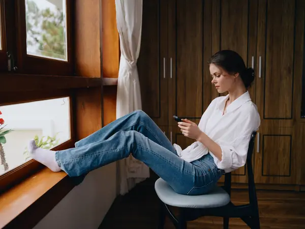 Ung Kvinna Sitter Vid Fönstret Med Telefon Handen Leende Tittar — Stockfoto