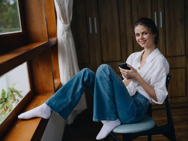 Νεαρή Γυναίκα Κάθεται Δίπλα Στο Παράθυρο Τηλέφωνο Στο Χέρι Χαμόγελο — Φωτογραφία Αρχείου
