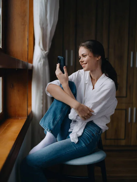 Mujer Joven Sentada Ventana Una Silla Con Teléfono Mano Sonriendo —  Fotos de Stock