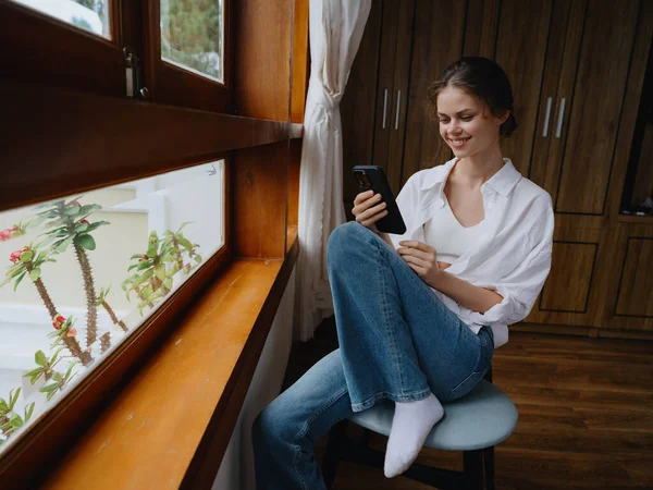 漂亮的女人 手里拿着电话 坐在窗前 木结构 舒适的生活 网上工作 舒适的氛围和美观 高质量的照片 — 图库照片