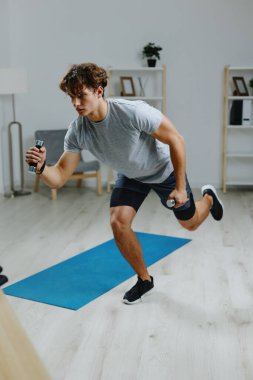 Yetişkin bir adam fiziksel olarak genç yaşam tarzı paspası kullanıyor. Gri spor kıyafetlerini esnetiyor. Spor halterleri takıyor.