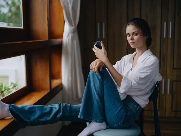 Νεαρή Γυναίκα Κάθεται Δίπλα Στο Παράθυρο Τηλέφωνο Στο Χέρι Χαμόγελο — Φωτογραφία Αρχείου