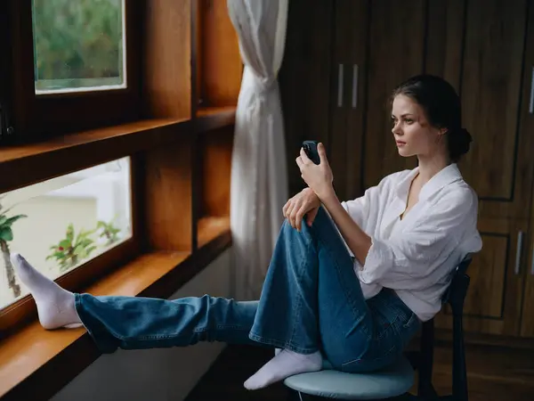 Young woman sitting by the window with phone in hand smile looking online, spring mood, home simple clothes, home cozy atmosphere, aesthetic lifestyle. High quality photo