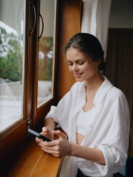 漂亮的女人 手里拿着手机 站在家里的窗口 自由职业者在度假 家里方便的生活方式与在线工作 网络安全的概念 聪明的家 高质量的照片 — 图库照片