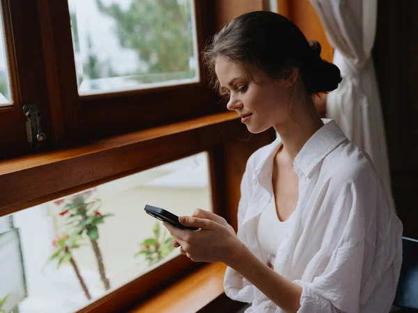 Beautiful woman with phone in hand sitting at the window with wooden frame of the house, home comfortable lifestyle with online work, cozy atmosphere and aesthetics, spring time. High quality photo