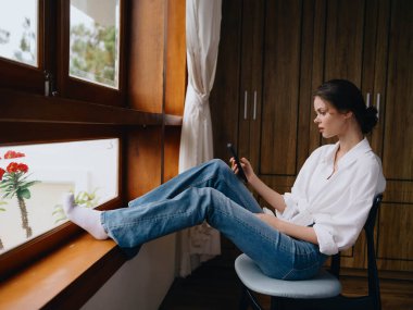 Pencerenin kenarında oturan genç bir kadın, elinde telefonla gülümsüyor çevrimiçi bakıyor, bahar havası, ev kıyafetleri, rahat atmosfer, estetik yaşam tarzı. Yüksek kalite fotoğraf