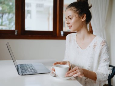 Evde rahat bir yerde, bir bardak içkiyle masa başında çalışan yaratıcı genç bir kadın. İnternette gülümsemesi, iletişimi ve eğitimi olan bir ofis çalışanı. Yüksek kalite fotoğraf