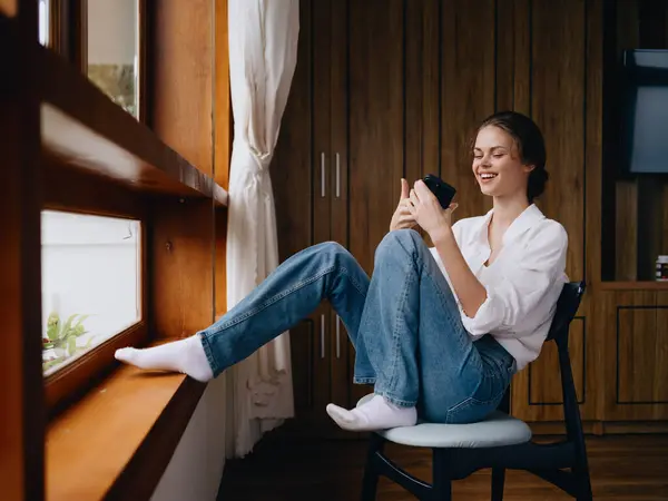 Jonge Vrouw Zit Bij Het Raam Met Telefoon Hand Glimlach — Stockfoto