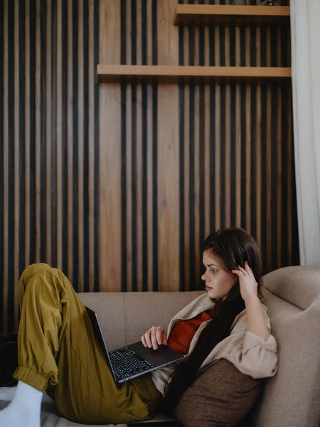 A woman lies on a couch with a laptop and stares pensively into the screen training a freelancer at work, a real lifestyle without retouching. High quality photo