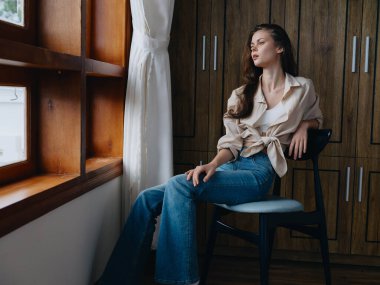 Pencerenin yanındaki bir sandalyede oturan bir kadın, rahat ve rahat, hafta sonları yaşam tarzı rahatlığı, güzel bir kızın düşünceli bakışları. Yüksek kalite fotoğraf