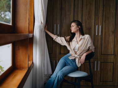 Pencerenin yanındaki bir sandalyede oturan bir kadın, rahat ve rahat, hafta sonları yaşam tarzı rahatlığı, güzel bir kızın düşünceli bakışları. Yüksek kalite fotoğraf