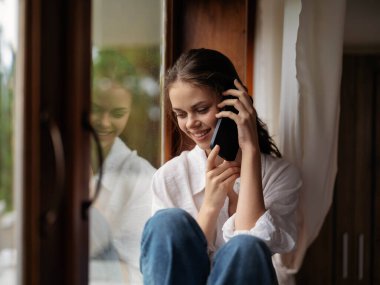 Pencerenin kenarındaki ahşap pencere pervazında oturan genç bir kadın elinde telefonla gülümseyen ve pencereden dışarı bakan bir kadın, sonbahar havası, rahat ev havası, estetik yaşam tarzı. Yüksek kalite fotoğraf