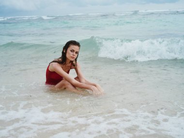 Kırmızı mayo giymiş bir kadın, Bali Adası sahilindeki dalgalarda okyanusun kenarında kumların üzerinde oturuyor ve gülümsüyor. Yüksek kalite fotoğraf