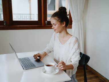 Evde rahat bir yerde, bir bardak içkiyle masa başında çalışan yaratıcı genç bir kadın. İnternette gülümsemesi, iletişimi ve eğitimi olan bir ofis çalışanı. Yüksek kalite fotoğraf