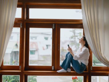 Pencerenin kenarındaki ahşap pencere pervazında oturan genç bir kadın elinde telefonla gülümseyen ve pencereden dışarı bakan bir kadın, sonbahar havası, rahat ev havası, estetik yaşam tarzı. Yüksek kalite fotoğraf