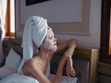 Genç bir kadın, duştan sonra kafasında nemlendirici yüz maskesi ve beyaz havluyla yatakta uzanıyor. Yaşam tarzı, cilt bakımı, gülümseme. Yüksek kalite fotoğraf