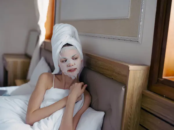 年轻女人躺在床上 洗完澡 脸上戴着保湿美容面罩 头上戴着白毛巾 生活方式家居护肤 面带微笑 高质量的照片 — 图库照片