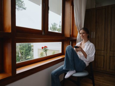 Elinde telefonla pencerede oturan güzel bir kadın, evin ahşap çerçevesi, çevrimiçi işlerle dolu rahat bir yaşam tarzı, rahat bir atmosfer ve estetik, bahar zamanı. Yüksek kalite fotoğraf