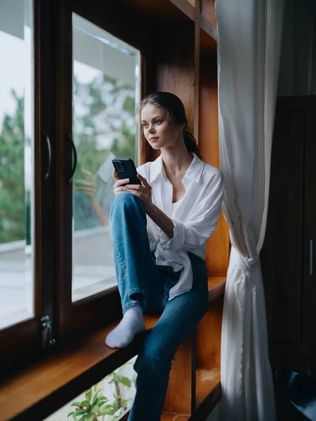 Young woman sitting on a wooden window sill by the window with a phone in hand smiling and looking out the window, autumn mood, home cozy atmosphere, aesthetic lifestyle. High quality photo