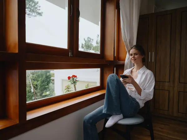 漂亮的女人 手里拿着电话 坐在窗前 木结构 舒适的生活 网上工作 舒适的氛围和美观 高质量的照片 — 图库照片