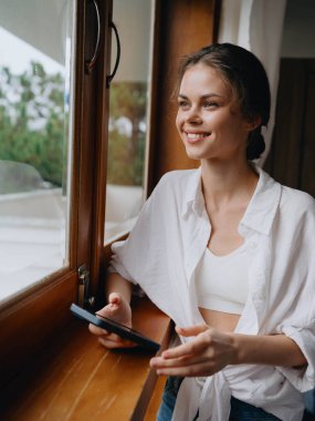 Ev penceresinde elinde telefonla bekleyen güzel bir kadın, tatilde serbest çalışan, çevrimiçi işi olan rahat bir ev hayatı, siber güvenlik konsepti, akıllı bir ev. Yüksek kalite fotoğraf