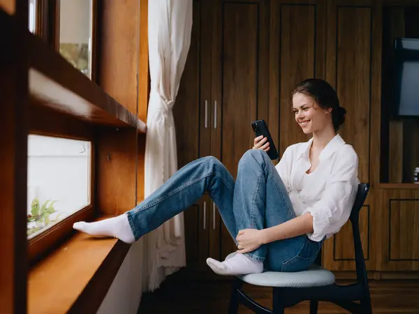 Pencerenin Kenarında Oturan Genç Bir Kadın Elinde Telefonla Gülümsüyor Çevrimiçi — Stok fotoğraf